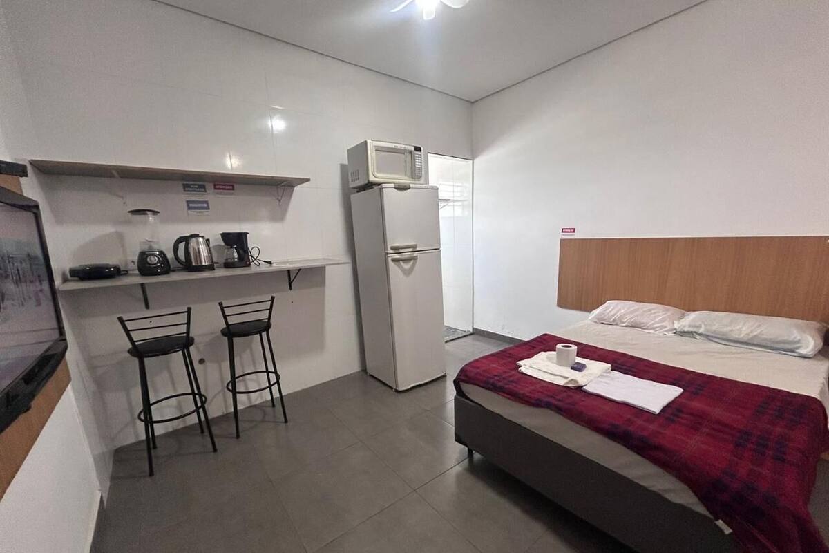 A compact room with a bed, a small kitchenette with a microwave, a refrigerator, and two bar stools.