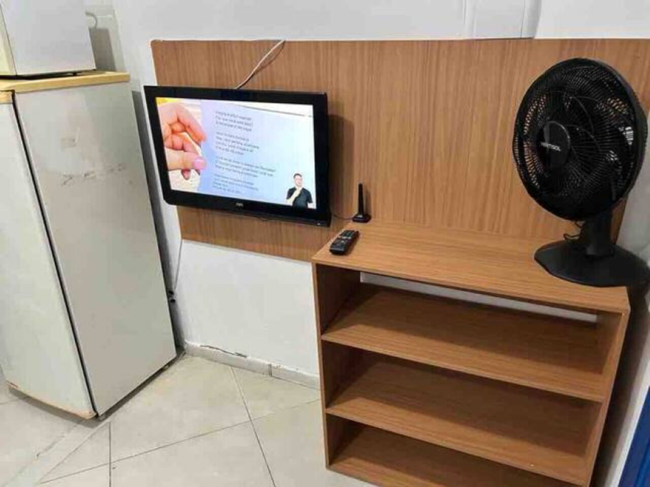 Quarto com televisão montada exibindo um vídeo, controle remoto e um ventilador.