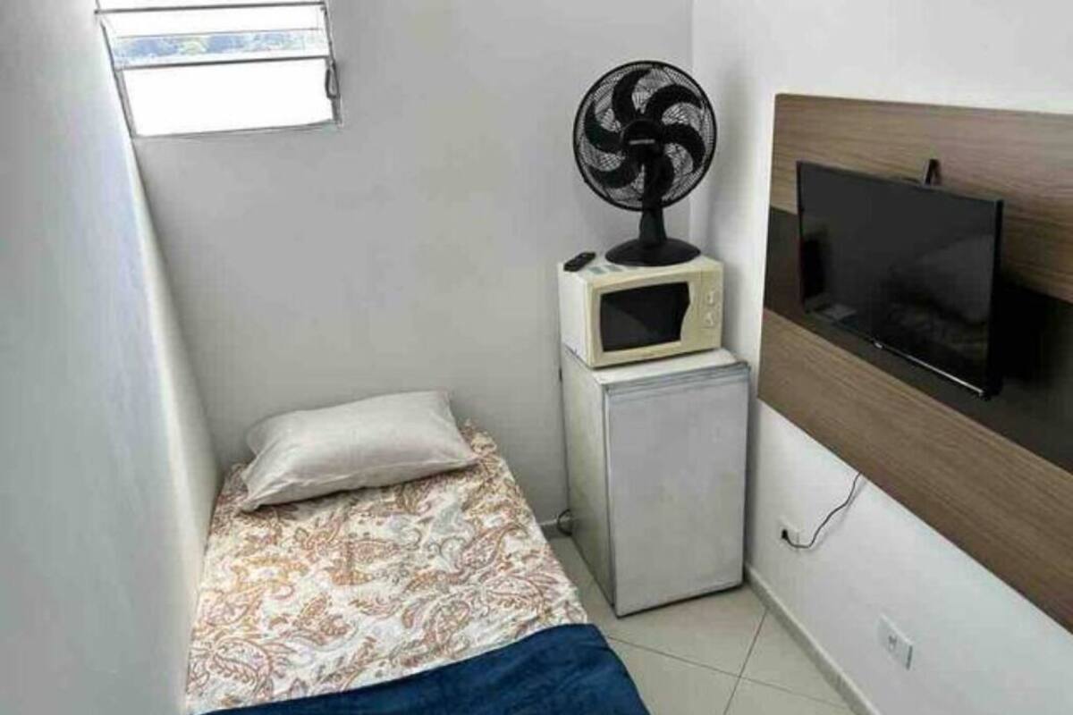 A small bedroom with a bed, a nightstand with a microwave, a fan, and a TV mounted on the wall.