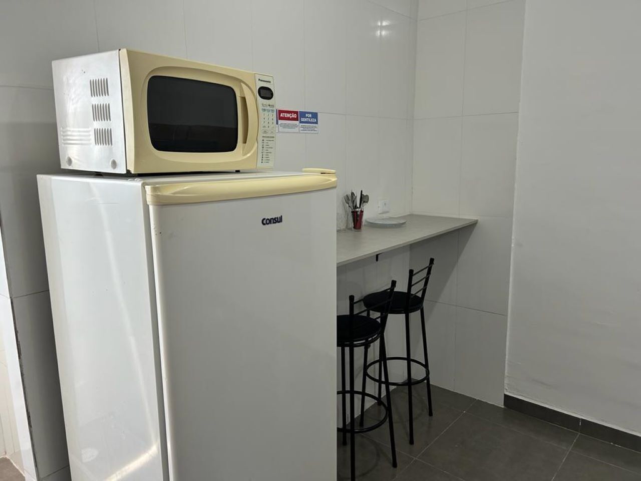 Eine kleine Küchenzeile mit einer Mikrowelle auf einem weißen Kühlschrank, einer kleinen Arbeitsplatte und zwei schwarzen Stühlen.