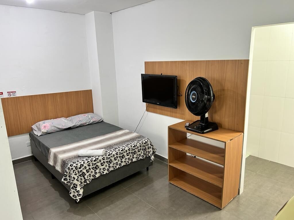 Un dormitorio con una cama, un televisor montado en la pared y un ventilador en un estante de madera.
