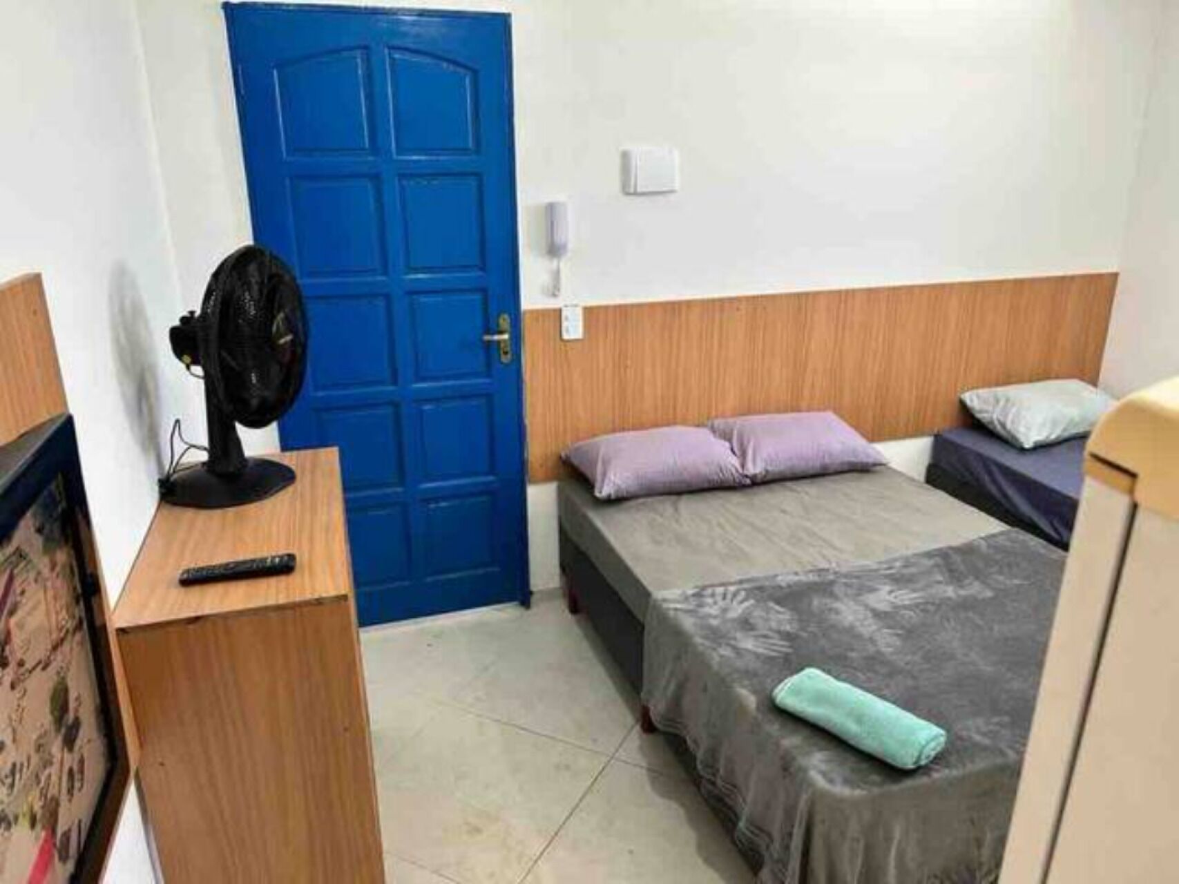 Una habitación con puerta azul, una cama con colcha gris, un escritorio de madera con ventilador y una mesita con control remoto.