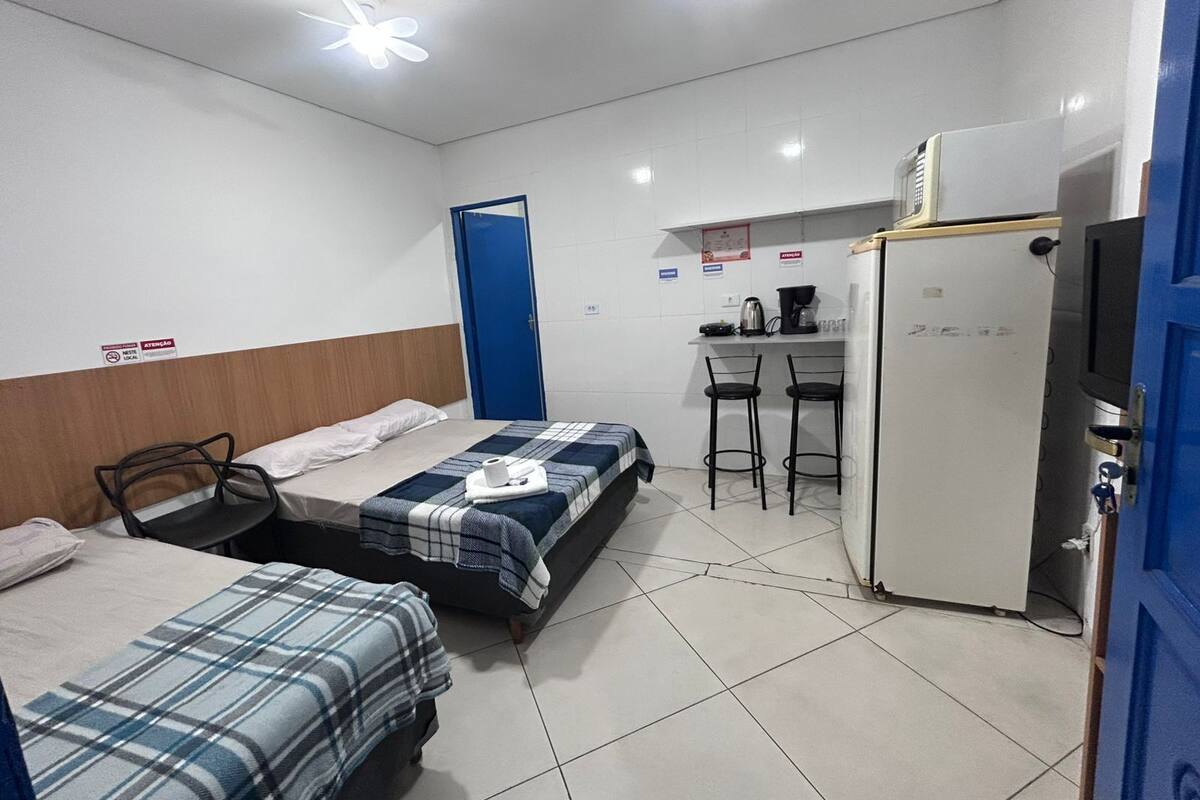 A small, clean room with two beds, a desk, a chair, a refrigerator, and a blue door.