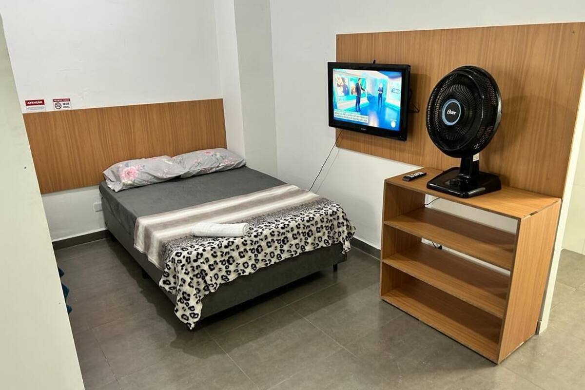 A hotel room with a bed, a TV mounted on the wall, and a standing fan.