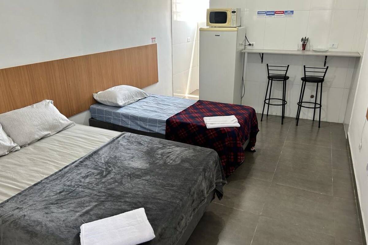 A dormitory room with three beds, a kitchenette, and two bar stools.