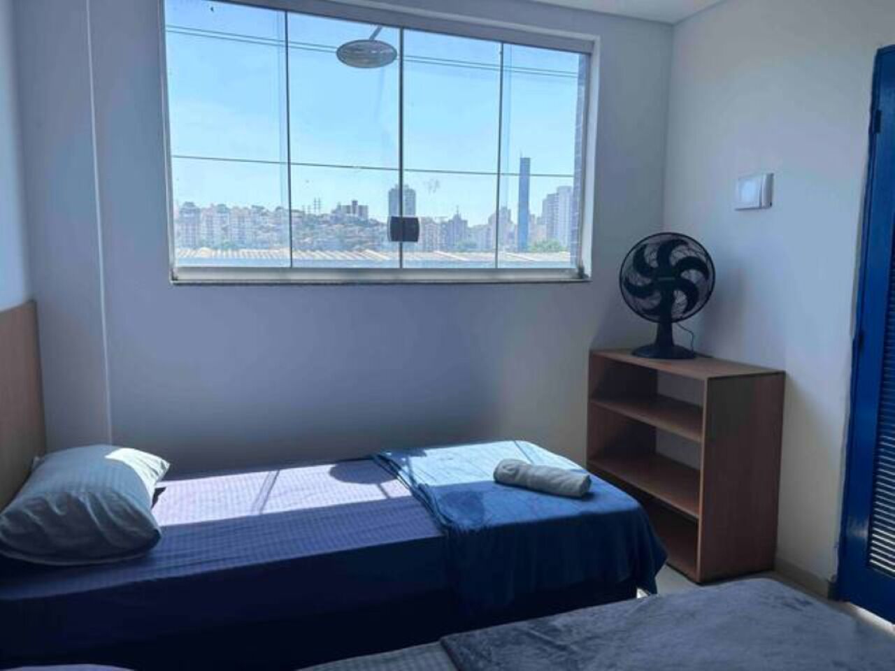 A small, single-bed room with a city view through a large window, a standing fan, and a wooden shelf unit.