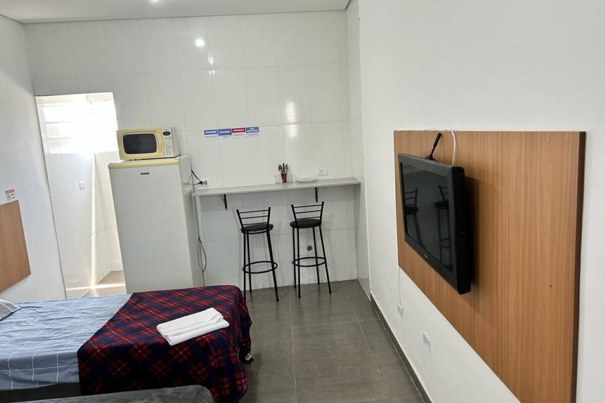 A small, compact room with two beds, a kitchenette, a TV mounted on the wall, and a window providing natural light.
