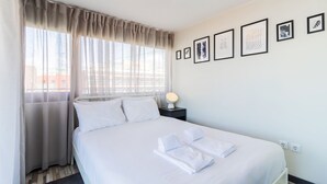 Hotellihuone, jossa on sänky, yöpöytä lampun kanssa ja ikkuna, jossa on läpikuultavat verhot.
