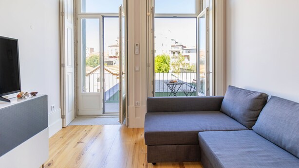 Un salon moderne avec un canapé gris, un téléviseur à écran plat posé sur un meuble blanc, et un balcon équipé d’une table et de chaises.