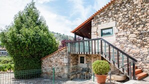 Ein Steinhaus mit einer Treppe, die zu einem Balkon führt, umgeben von Grünpflanzen und einem Steinzaun.