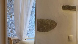 Une chambre avec un mur en pierre, un lit avec des rideaux blancs, une table de chevet en bois et une petite table surmontée d’un chemin de table.