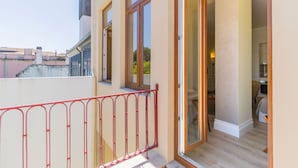 Ein Balkon mit roter Metallreling, eine Holztür zu einem Innenraum mit Esstisch und Stühlen sowie Blick auf benachbarte Gebäude.