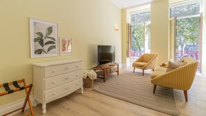 Una sala de estar con televisor, sofá, sillón, cómoda y ventana con vista a árboles.