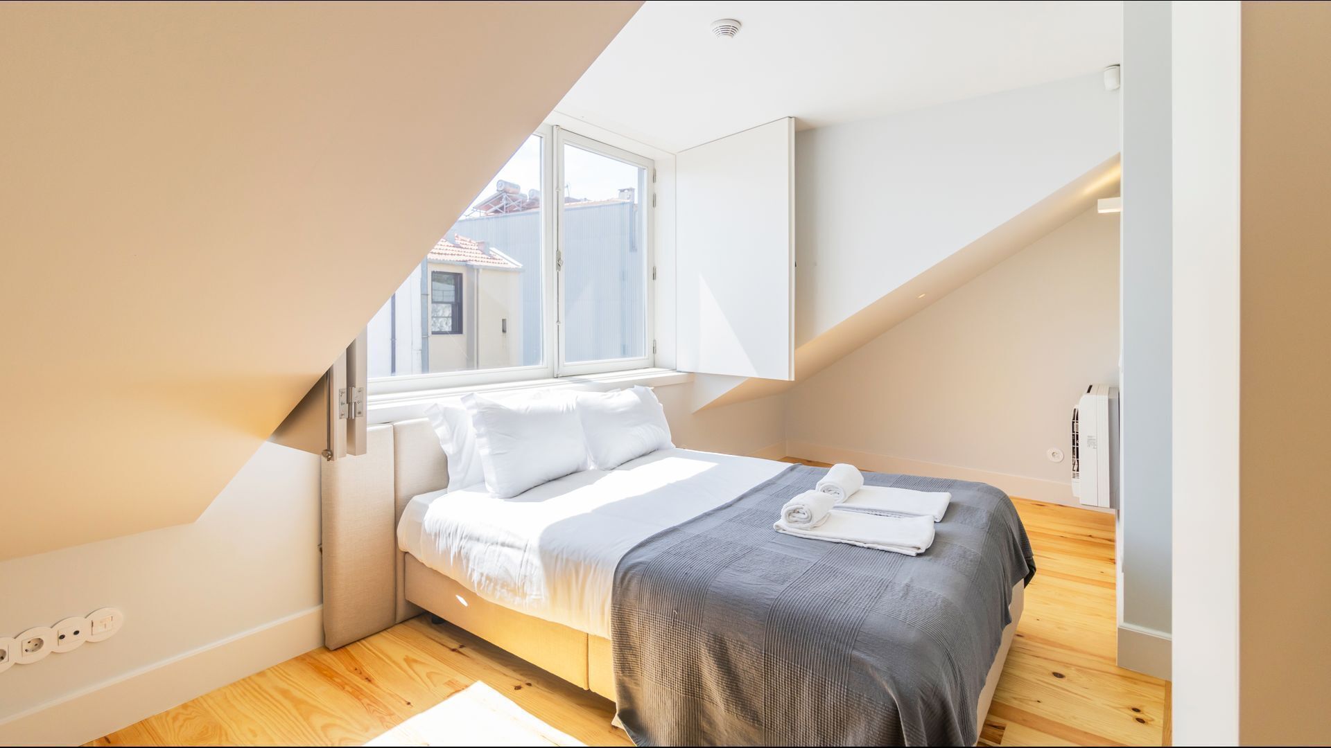 Un dormitorio con techo inclinado, una cama con ropa de cama blanca, un edredón gris y una ventana con vistas a edificios.