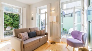 Una sala moderna con un sofá color beige, un sillón morado y una lámpara de pie. Amplias ventanas ofrecen luz natural y vistas al exterior.