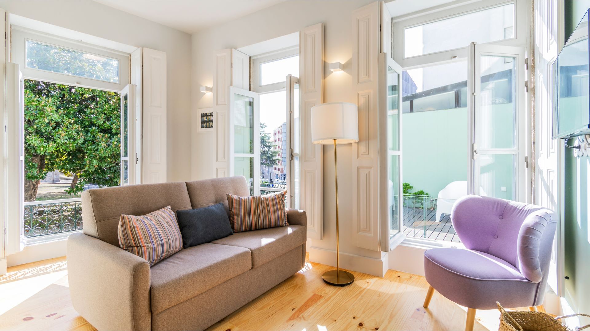 Un soggiorno moderno con un divano color beige, una poltrona viola e una lampada da terra. Ampie finestre offrono luce naturale e vista sull'esterno.
