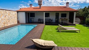 Una piscina con cubierta de madera, un césped con dos sillas de jardín y un muro de piedra.