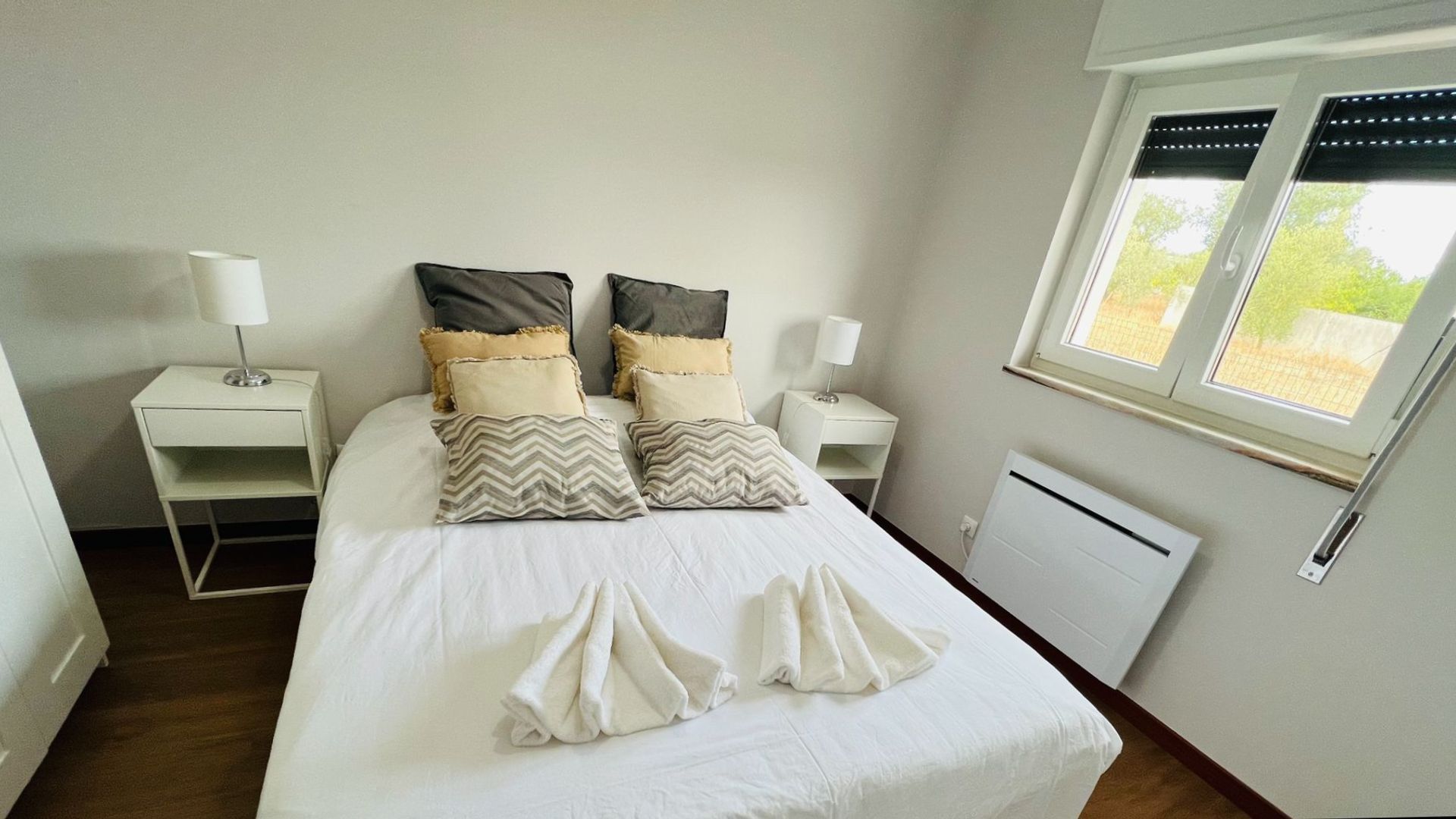 A neatly made bed with white bedding and decorative pillows, a bedside table with a lamp, and a window with a view of greenery.