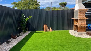 Un patio trasero cercado con césped artificial, una zona de parrillada de ladrillo y macetas decorativas.