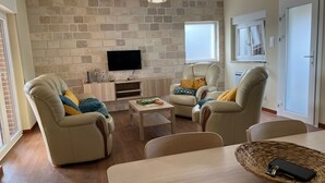 A living room with a stone wall, a flat-screen TV, two leather armchairs, a coffee table, and a dining table with a woven basket.