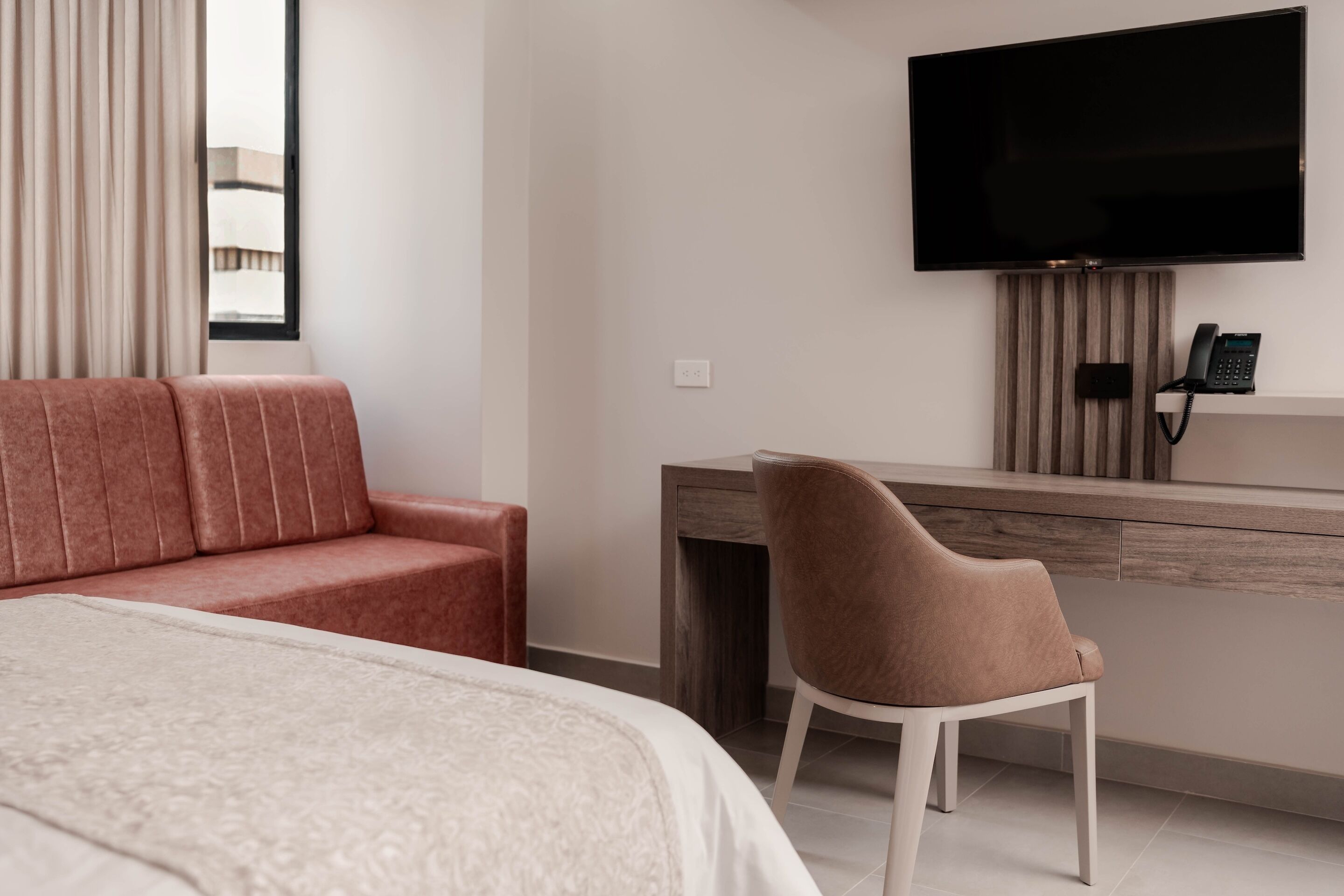 Quarto de hotel moderno com uma cama grande, um sofá vermelho, uma escrivaninha com cadeira, uma televisão de tela plana e um telefone.