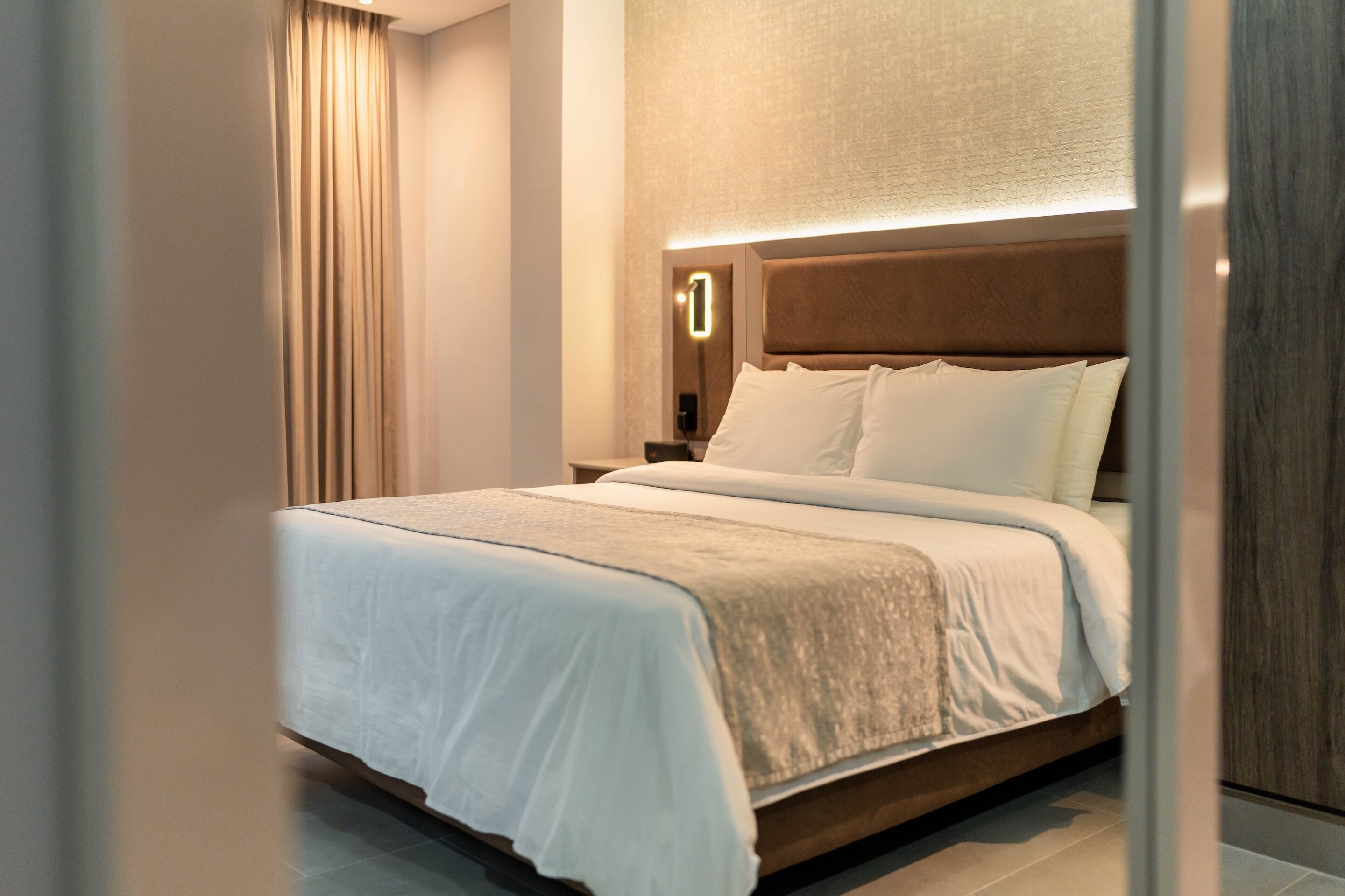 A modern hotel room with a large bed, a nightstand, a wall-mounted light, and a curtain.