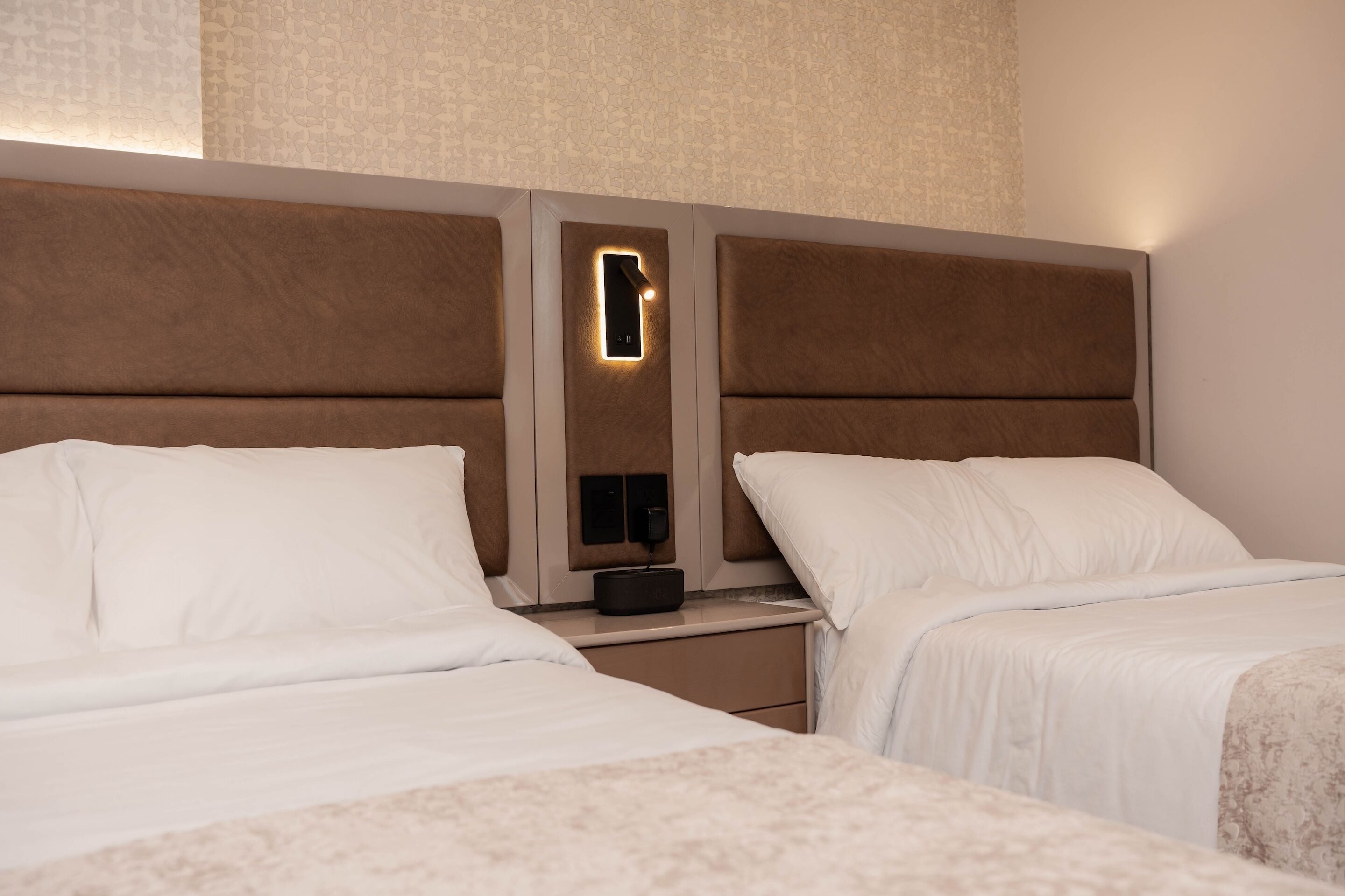 Habitación de hotel con dos camas, una mesita de noche con un teléfono y un pequeño dispositivo, y una pared con textura.