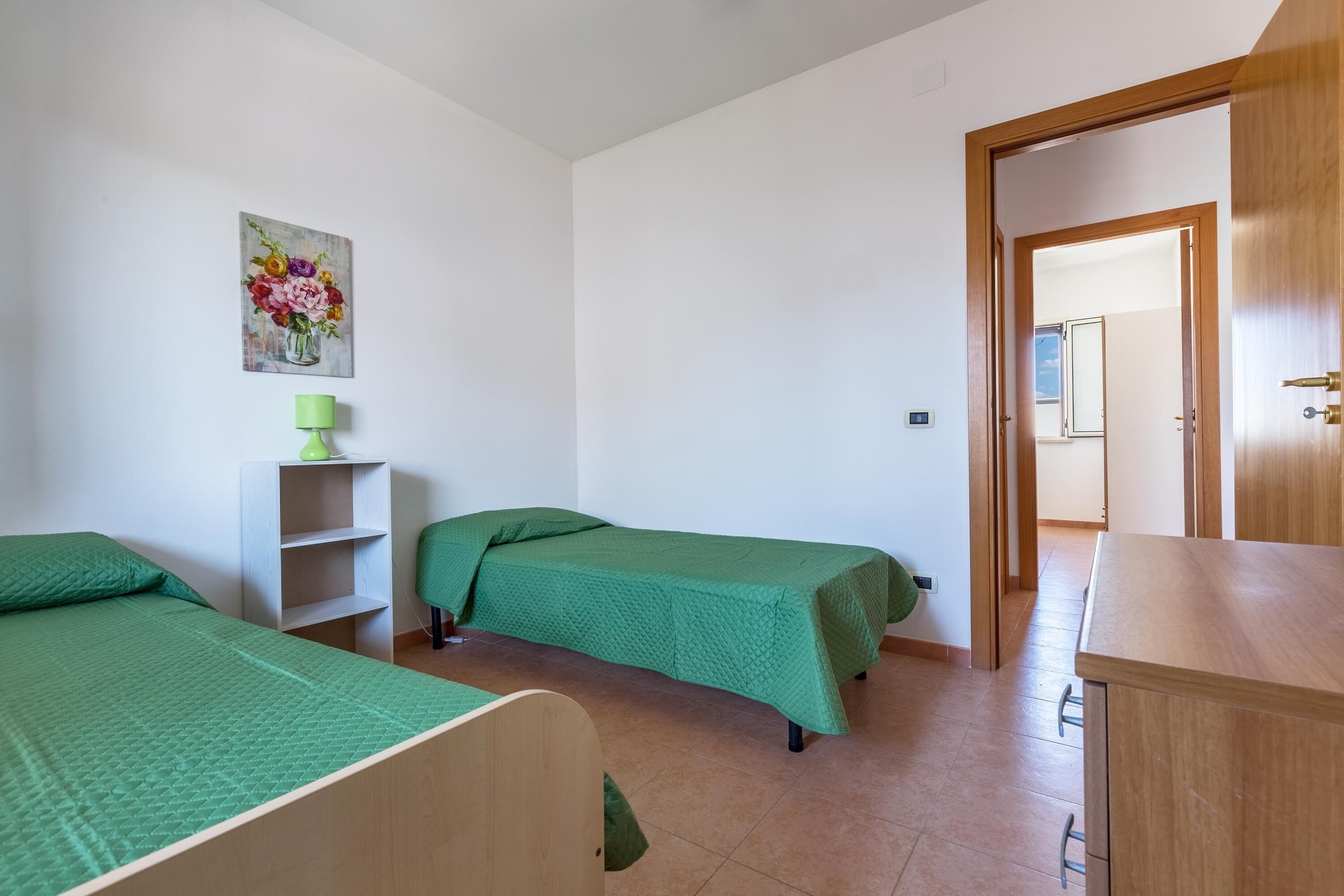 Una habitació amb dos llits individuals, una obra d'art mural verd, una petita estant blanca i una porta de fusta que dona a una altra habitació.