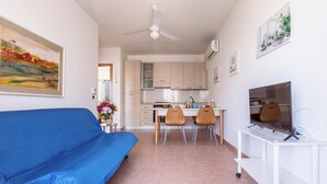 Ein modernes Wohnzimmer mit einem blauen Sofa, einem Essbereich mit zwei Stühlen, einer Küche mit hellen Schränken und einem Fernseher auf einem weißen Schrank.