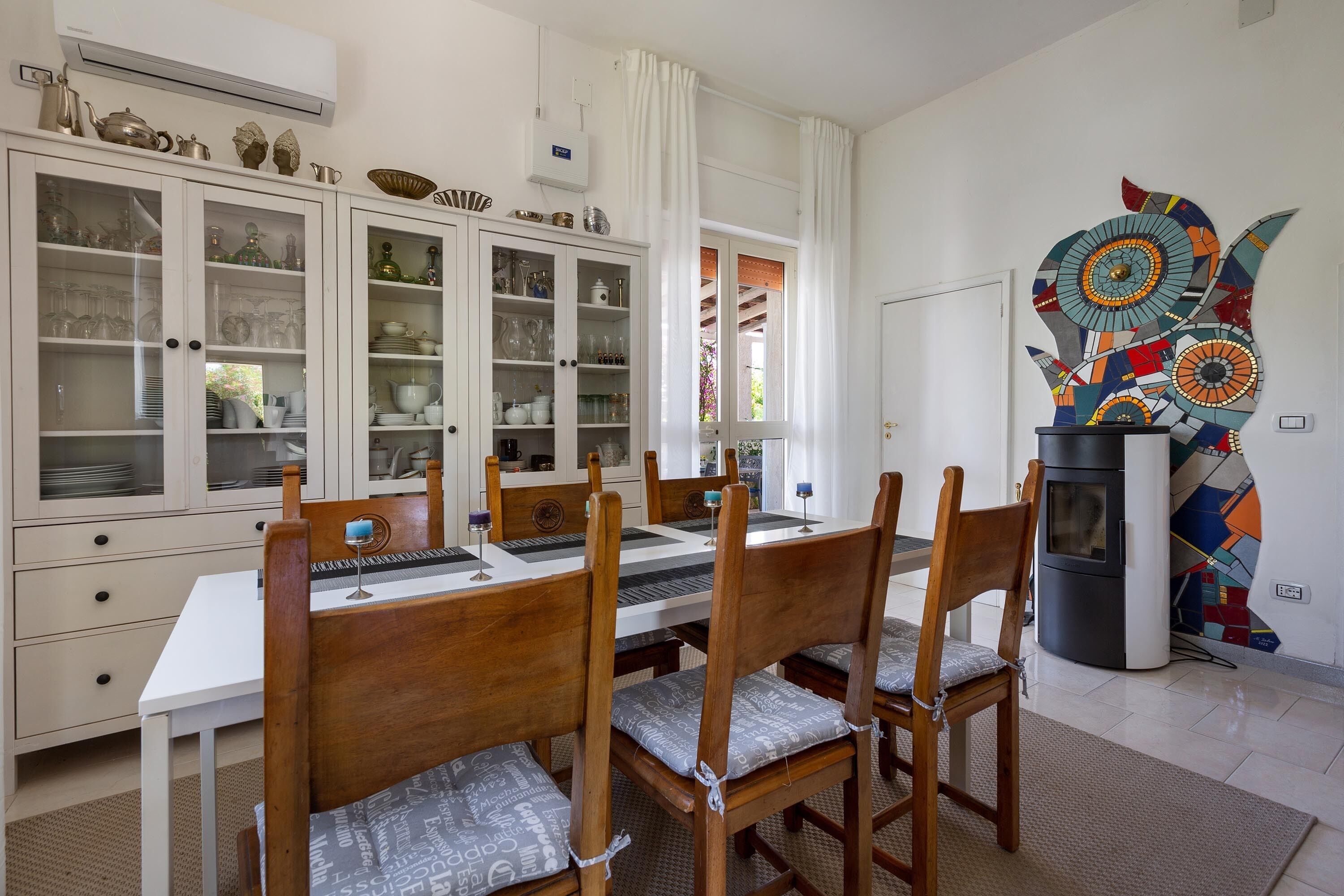 A dining area with a white table and wooden chairs, a cabinet with glass doors, and a colorful abstract art piece on the wall.