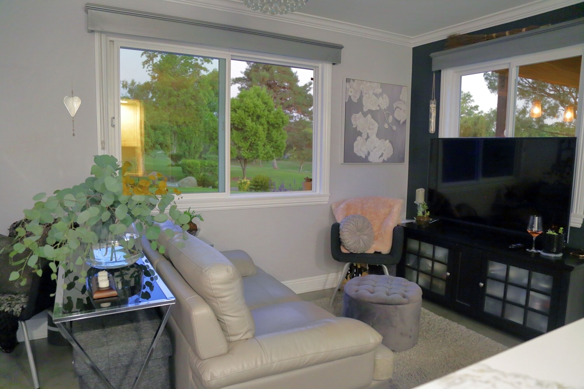 A living room with a beige sofa, a glass coffee table, a TV on a black stand, and a window with a view of greenery.