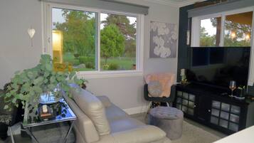 A living room with a beige sofa, a glass coffee table, a TV on a black stand, and a window with a view of greenery.