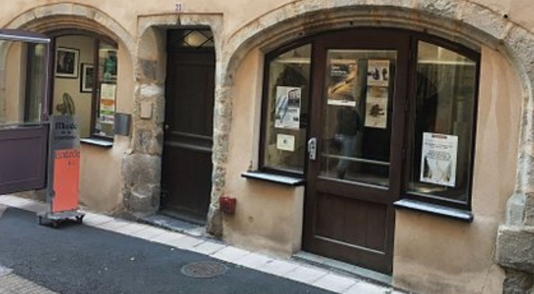 Un bâtiment doté d’une façade en pierre, de deux portes en arc de cercle et d’une petite fenêtre sur laquelle est affichée une affiche.