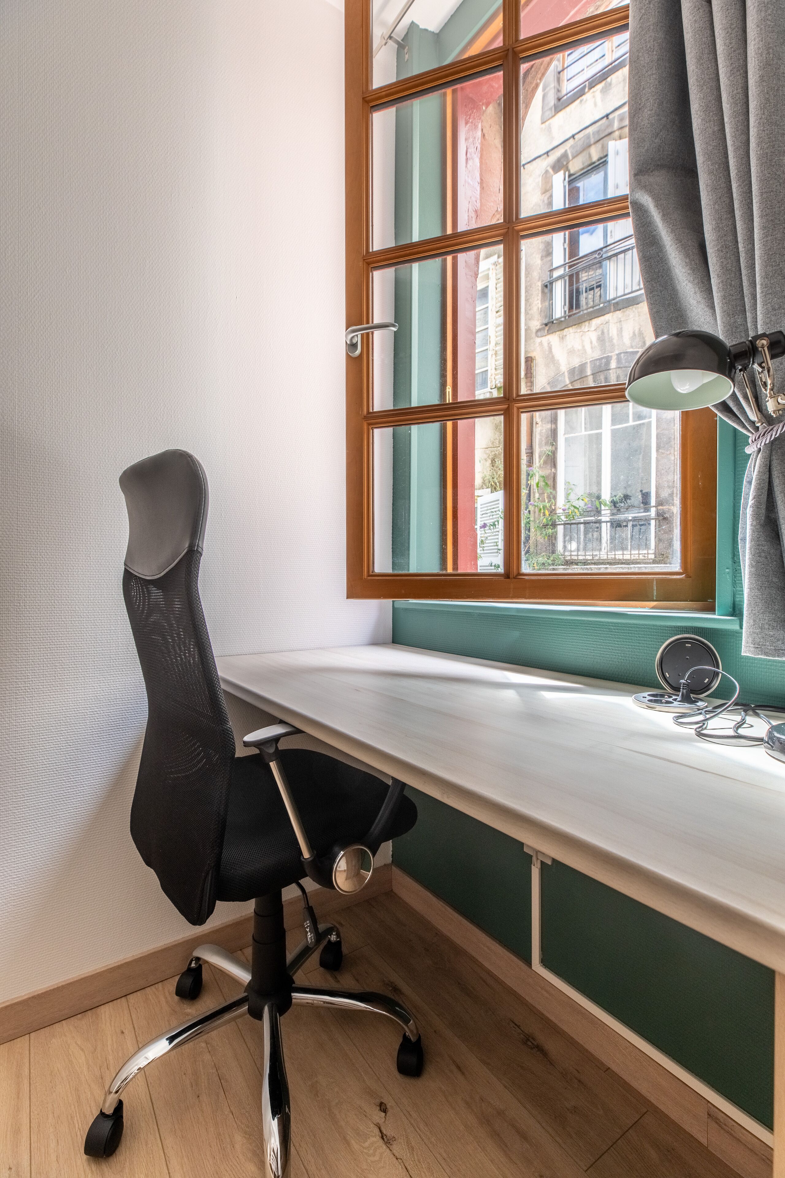 Een modern bureau met een zwarte bureaustoel, een raam met uitzicht op gebouwen, en een bureaulamp.