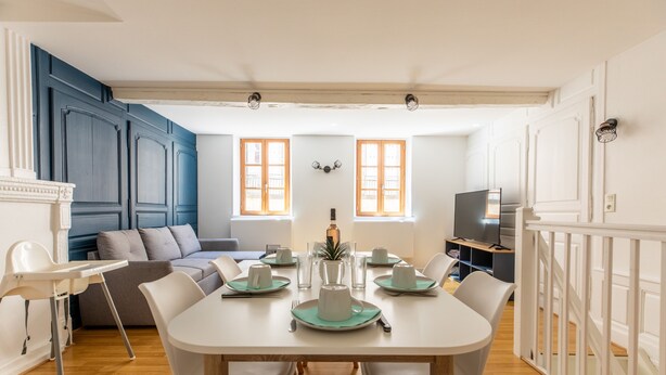 Un coin repas avec une table blanche dressée pour quatre personnes, entourée de chaises blanches. On aperçoit un canapé et un téléviseur au fond.