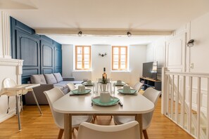 Un coin repas avec une table blanche dressée pour quatre personnes, entourée de chaises blanches. On aperçoit un canapé et un téléviseur au fond.