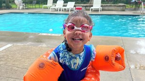 Un enfant se trouve dans un espace autour d’une piscine, avec des chaises longues et des arbres en arrière-plan.