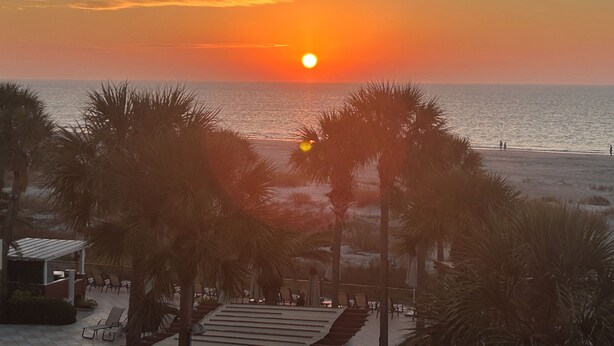 Une vue sur le coucher de soleil depuis un endroit situé au bord de la plage, avec des palmiers et un espace patio.