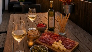 Un tavolo di legno con una bottiglia di vino, bicchieri, tagliere di salumi, olive e grissini su un balcone affacciato su un tramonto.