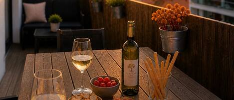 A wooden table with a wine bottle, glasses, charcuterie board, olives, and breadsticks on a balcony overlooking a sunset.