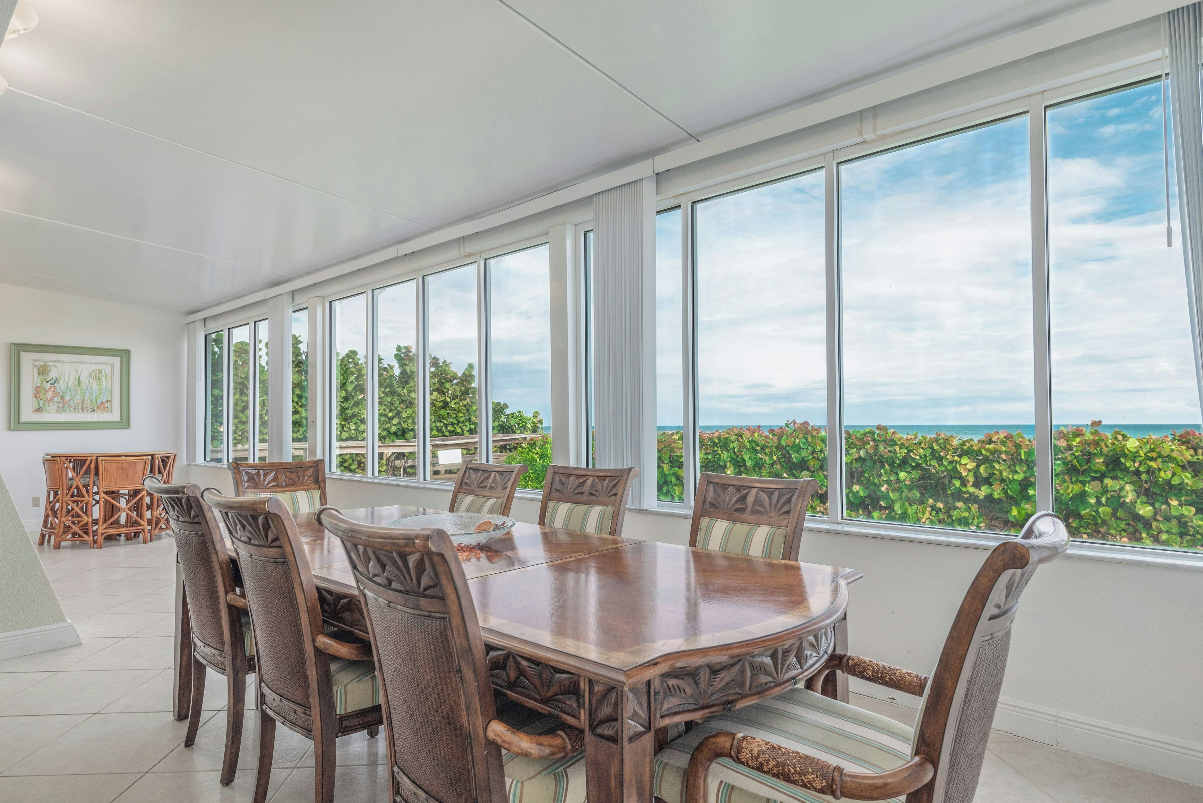 A dining area with a large table and chairs, large windows with a view of the ocean and greenery.
