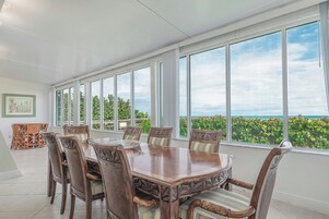 Ein Essbereich mit einem großen Tisch und Stühlen, große Fenster mit Blick auf das Meer und viel Grün.