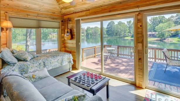 A cabin with a bed, sofa, and a table with a board game. Large windows offer views of a lake and deck furniture. - Kilgore Lakefront Home w/ Private Dock & Pier! (Kilgore)