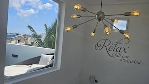 Una habitación con una lámpara moderna, un ventanal con vistas a palmeras y edificios, y una pared con el letrero 'Relax Chill out & Unwind'.