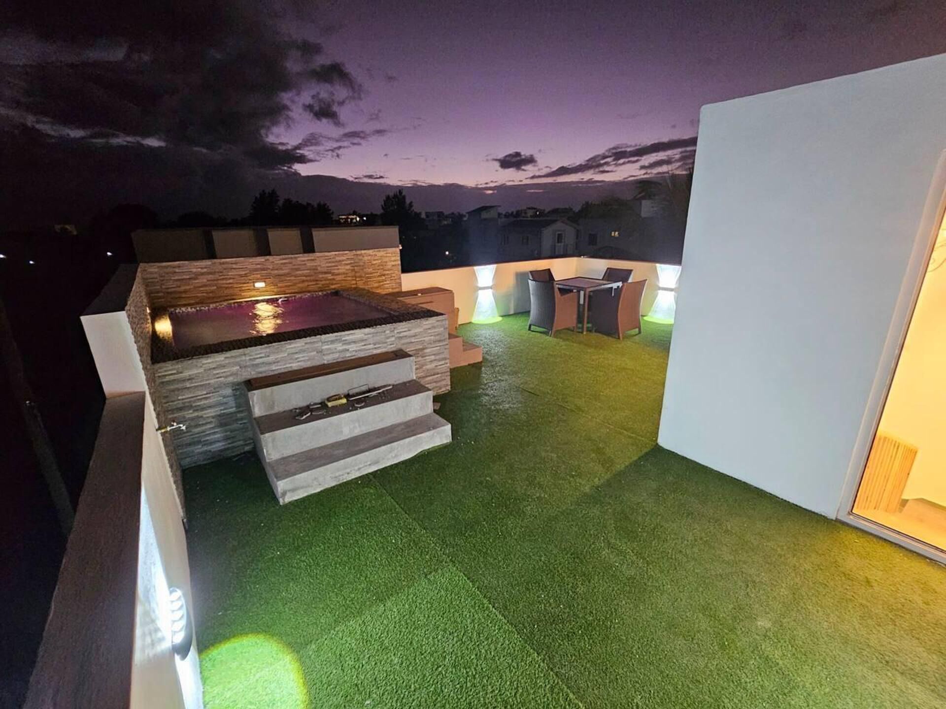 Una terraza en la azotea con un jacuzzi, un espacio de estar con mesa y sillas, y vistas del horizonte urbano al atardecer.