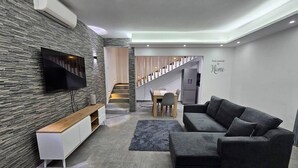 Una sala de estar moderna con un sofá modular, un televisor de pantalla plana montado en una pared con textura, una consola de madera, un comedor con mesa y sillas, y una escalera que sube a otro nivel.