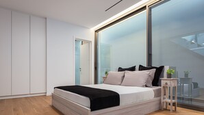 A modern bedroom with a bed, a small wooden nightstand, a glass wall, and a staircase visible through the glass. - Athenian Riviera Prime Residence  (Vari-Voula-Vouliagmeni)