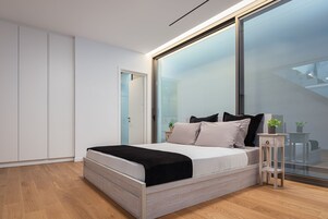 Quarto moderno com cama, mesa de cabeceira de madeira pequena, parede de vidro e escada visível pelo vidro.