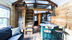 A cabin interior with a wooden staircase, a kitchen area, and a seating area with a sofa and bar stools. - ONYX OWL CABIN: Private Hot Tub⁘Fire Pit⁘Heated Patio⁘Vineyard⁘Hiking⁘Golf (Scaly Mountain)