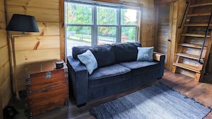 A cozy cabin interior with a wooden floor, a dark blue sofa with cushions, a wooden chest of drawers, a lamp, and a large window offering a view of greenery outside. - ONYX OWL CABIN: Private Hot Tub⁘Fire Pit⁘Heated Patio⁘Vineyard⁘Hiking⁘Golf (Scaly Mountain)